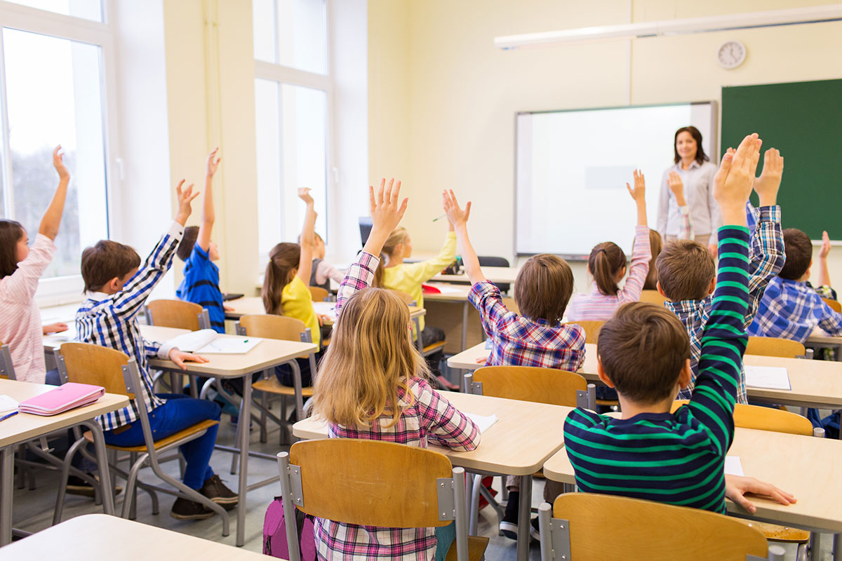 Students raising their hands in classroom with a teacher standing in front of the board